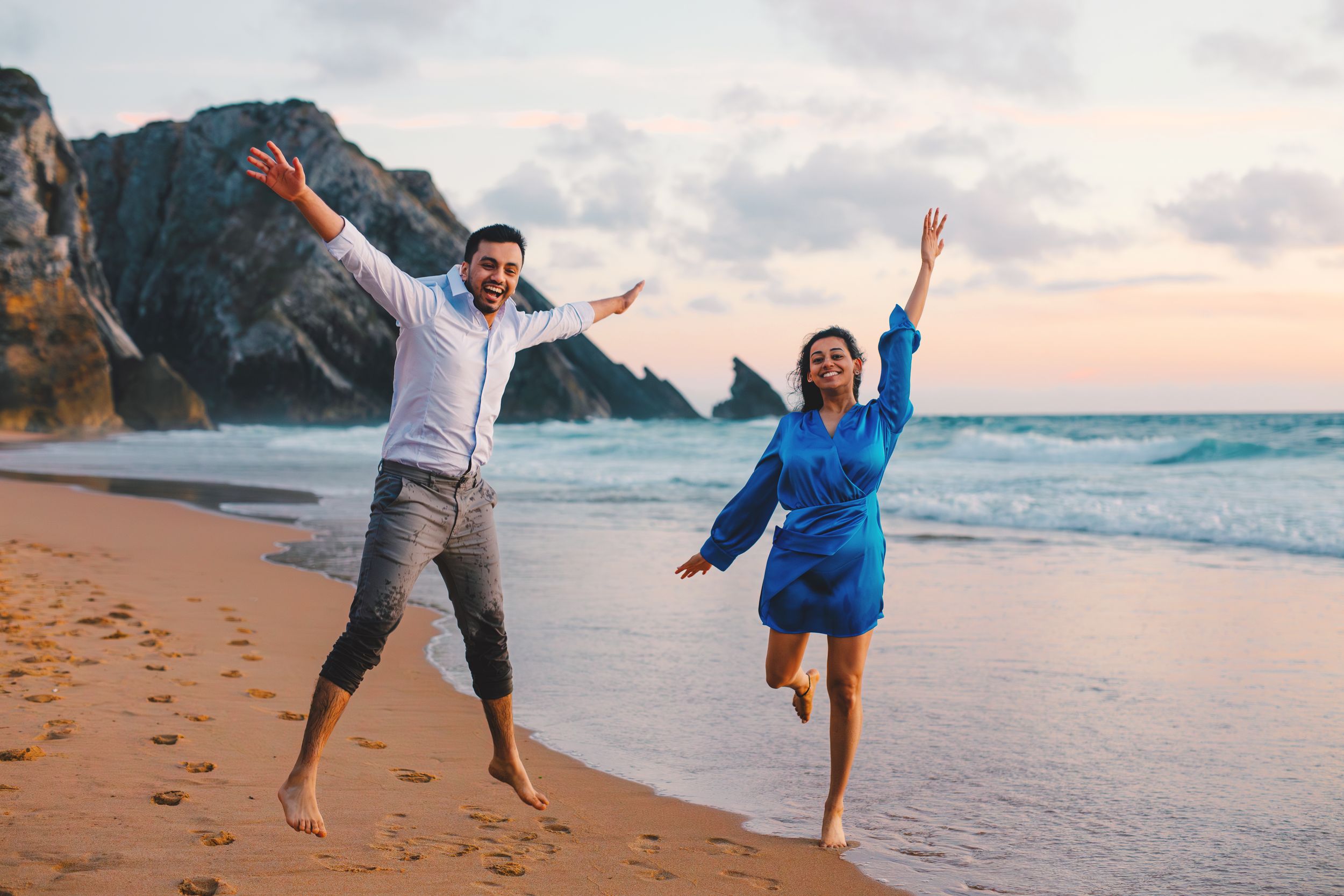 People enjoy a playful moment together on a sandy beach at sunset with waves and cliffs in the background.