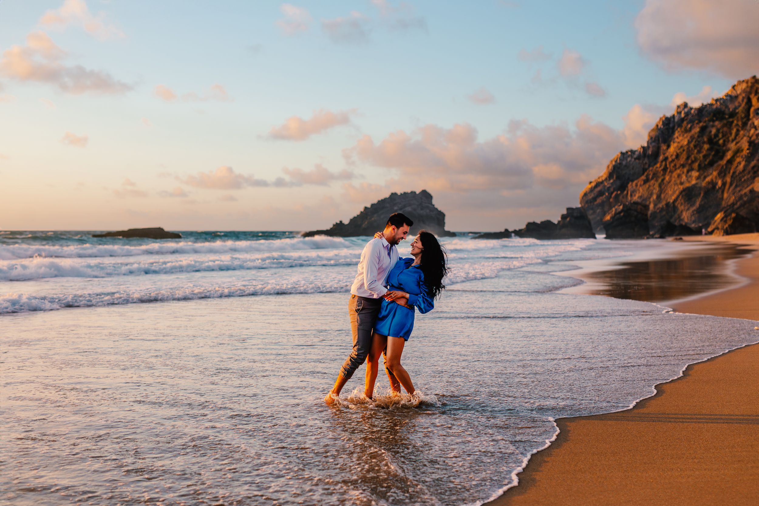 Couple walking and playing along a sandy beach at sunset with waves crashing on the shore.