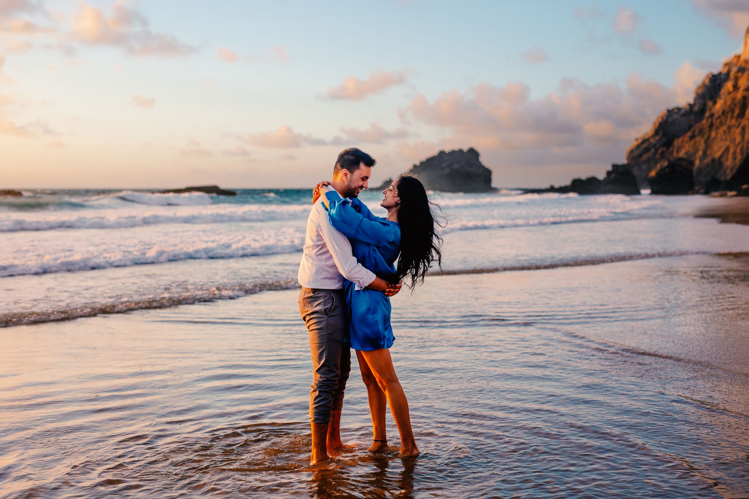 Couple sharing romantic moments at sunset on a beach with rocky cliffs and ocean waves in the background.