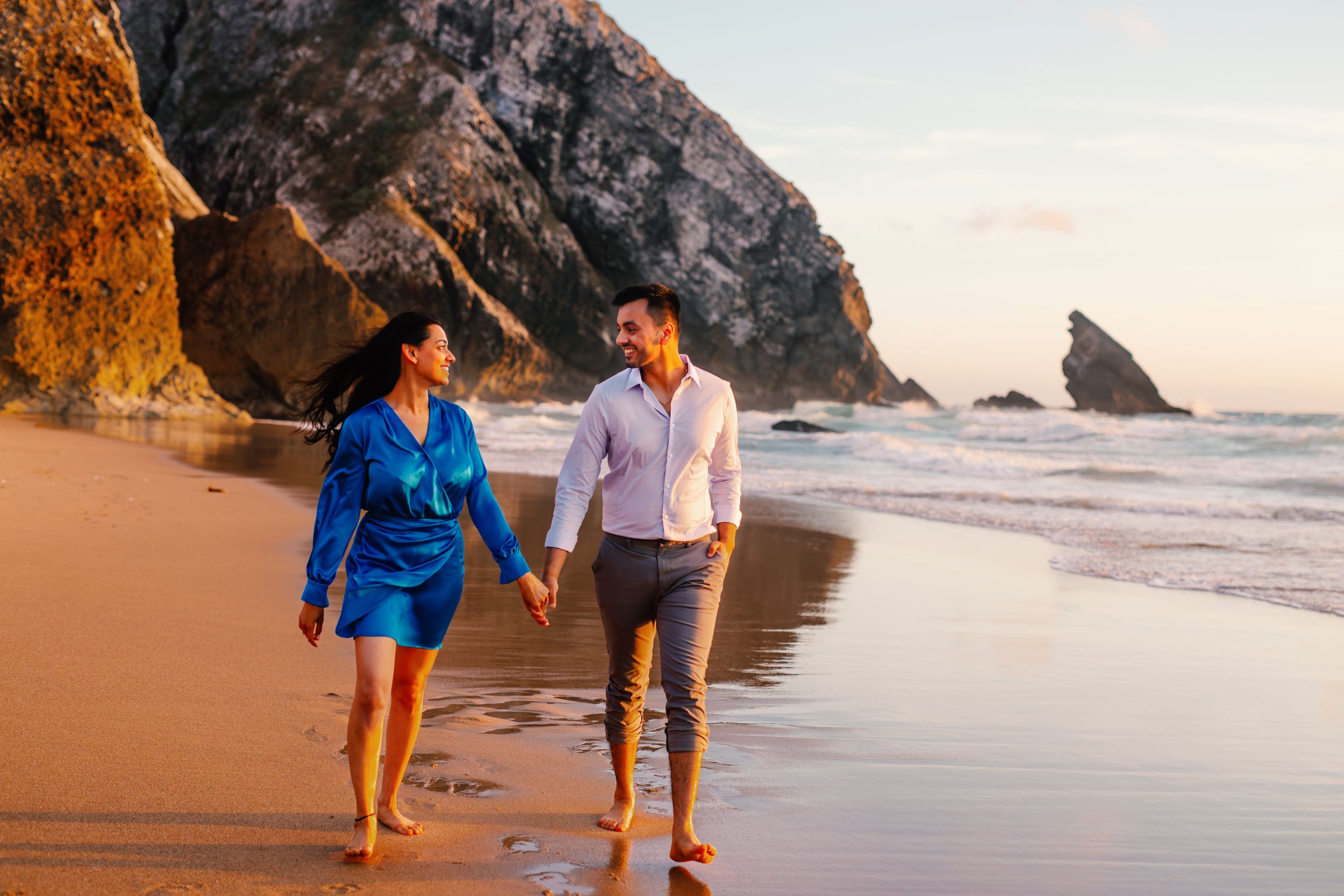 Couple walking hand in hand along a scenic beach at sunset with rocky cliffs in the background.