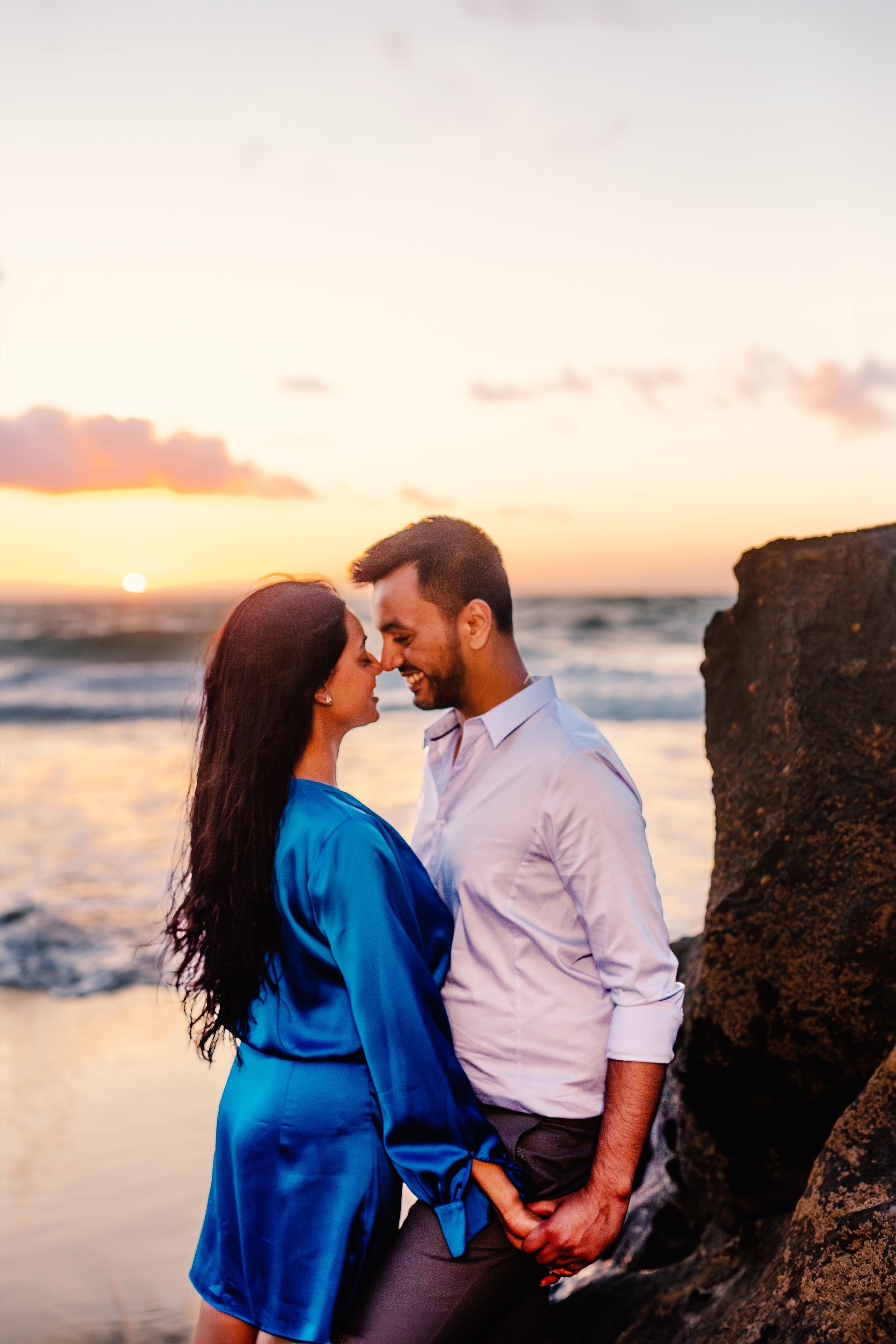 A couple embraces during sunset at a rocky beach, with the ocean and warm sky creating a romantic backdrop.