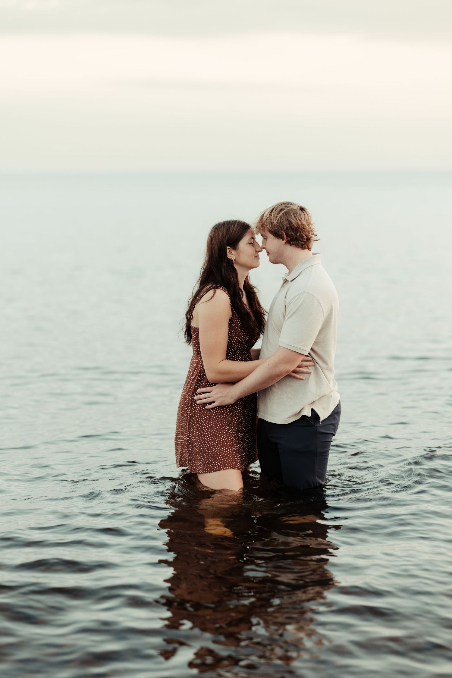 A romantic couple embraces while standing in calm ocean waters during sunset.