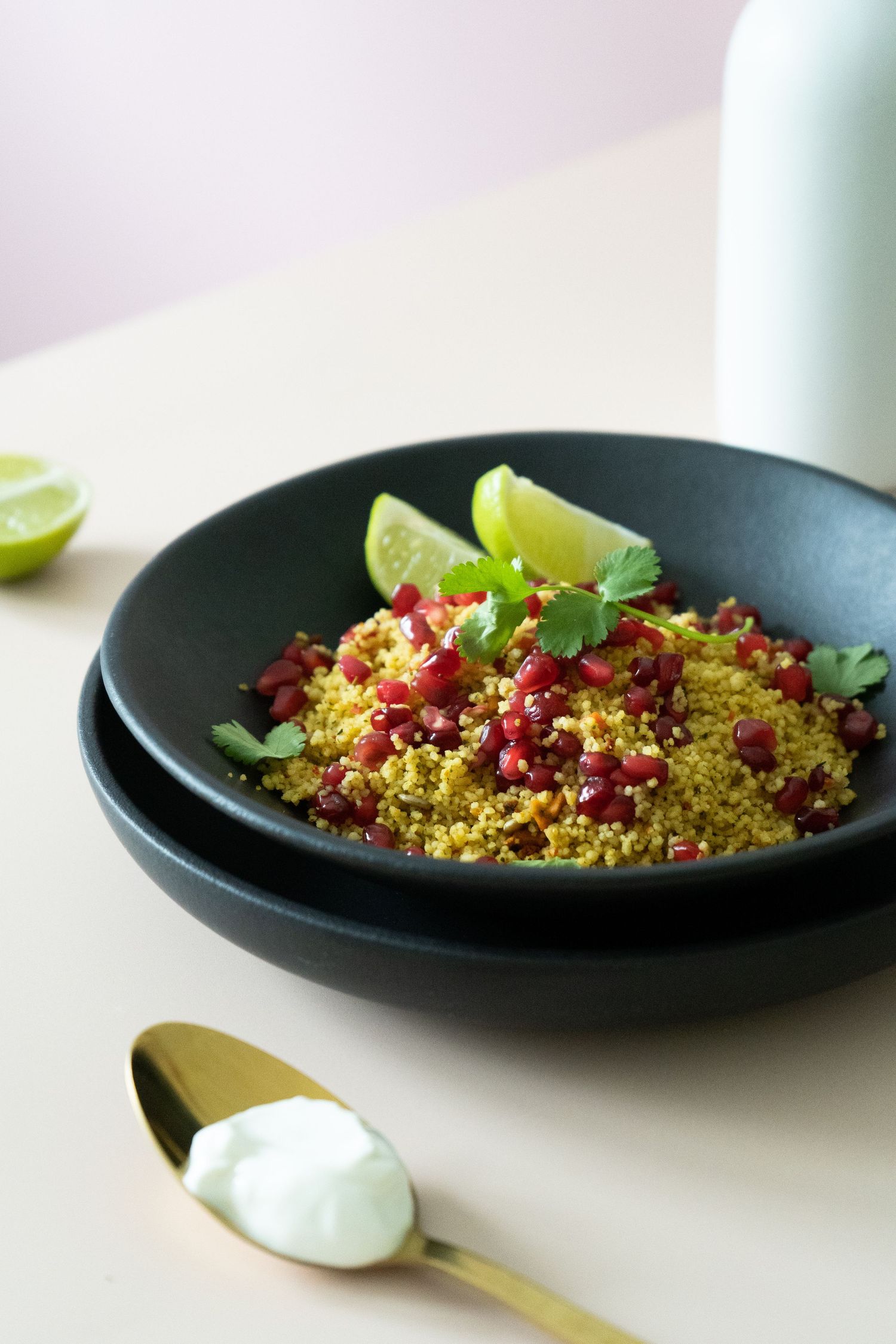 Modern lifestyle product photo of cous cous recipe in luxury tableware black bowl, topped with pomegranate seeds and yoghurt on side.