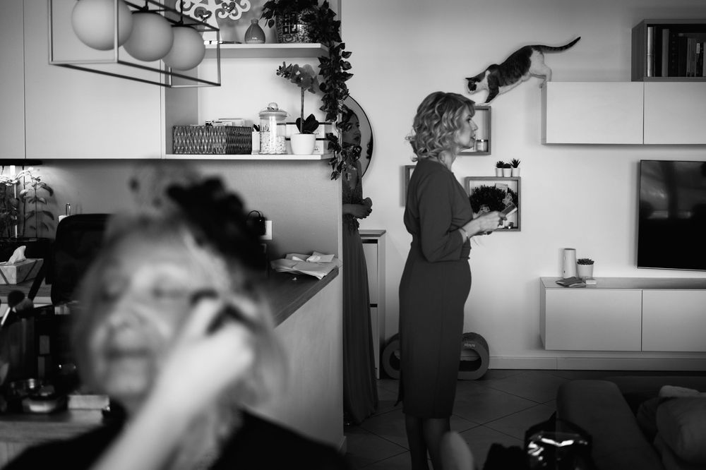 Black and white photo showing a figure in a dress reaching up to cabinets in a kitchen interior.