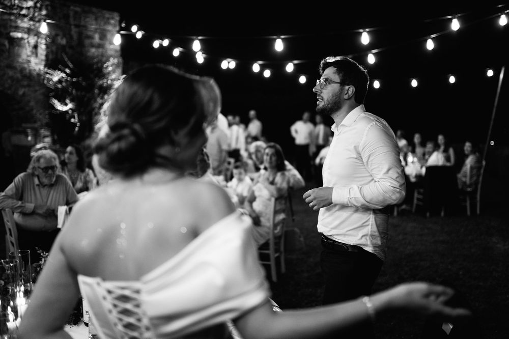 Wedding guests dance and celebrate on a dimly lit dance floor under hanging string lights at an evening reception.