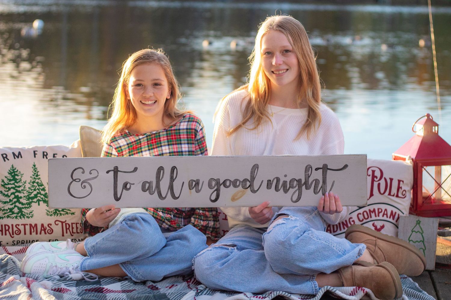 People holding Christmas-themed wooden signs while sitting outdoors near a lake at sunset.