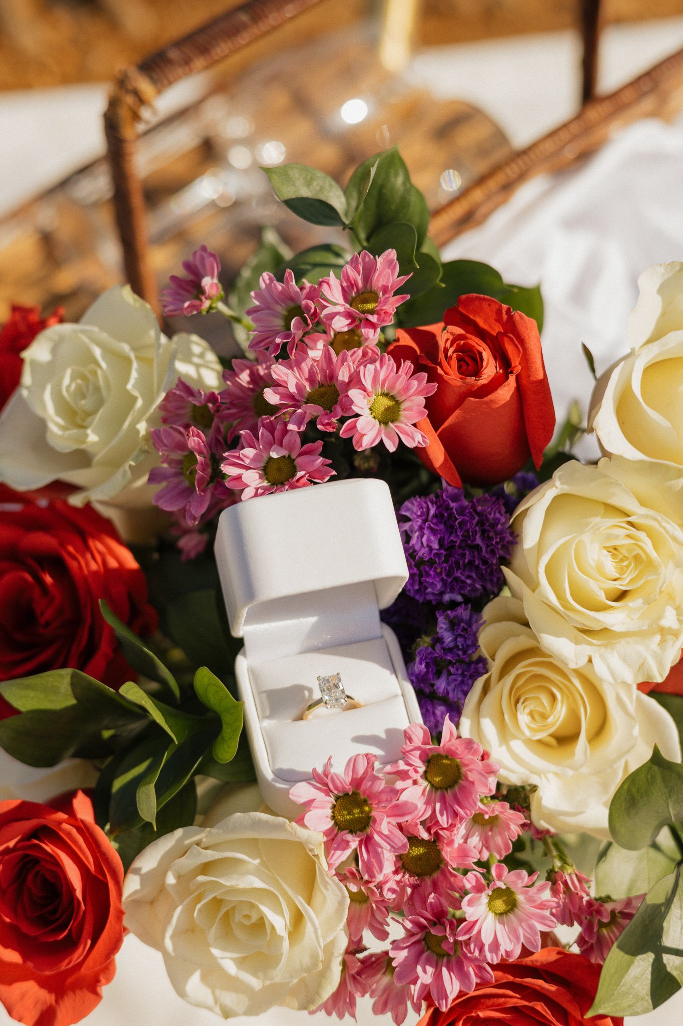 Diamond engagement ring in white box surrounded by colorful red, white, and pink roses and flowers.