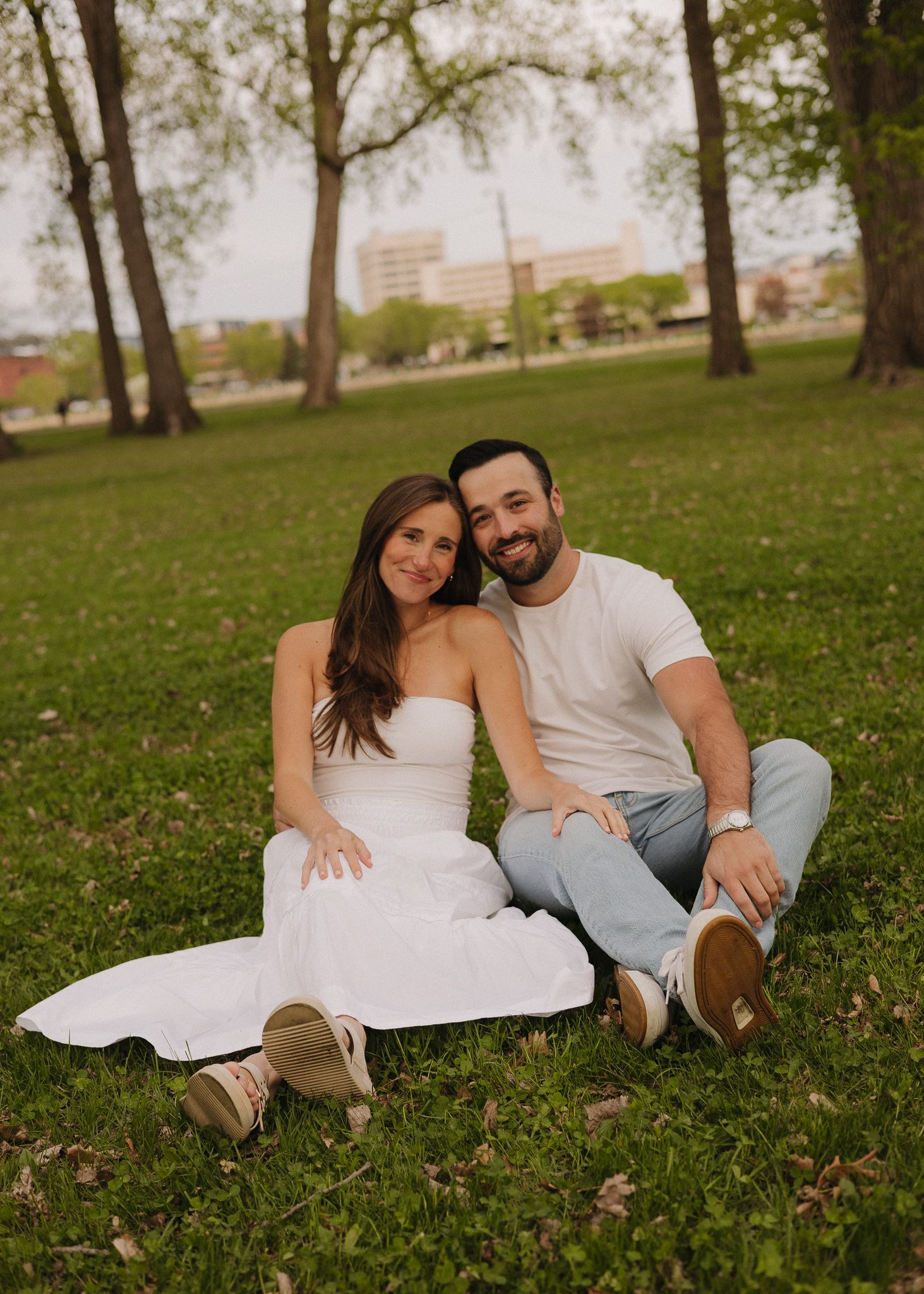 Happy couple sitting together on green grass in a park, wearing coordinated white outfits on a sunny day.