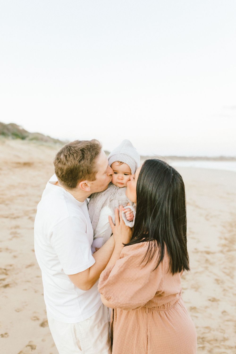 A loving family shares tender moments together on a sunlit beach in matching light-colored outfits.