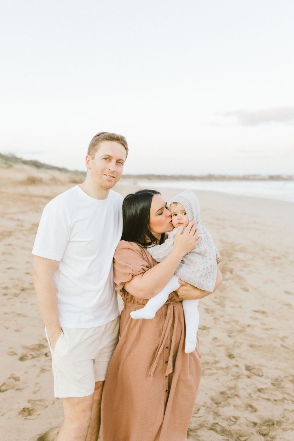 Family in white and tan outfits sharing tender moments together on a sandy beach at sunset.
