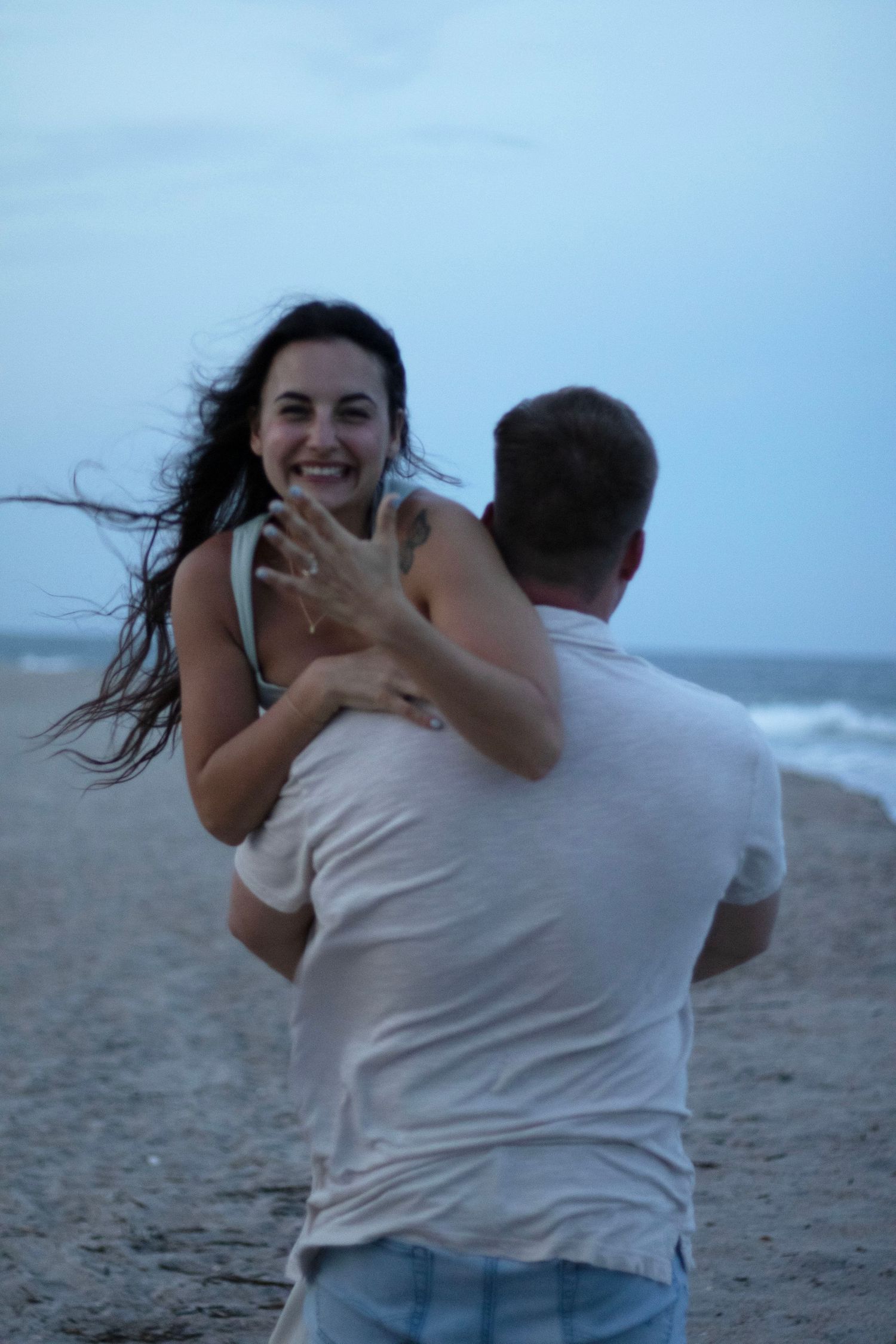 Joyful beachside engagement moment captured as couple celebrates by the ocean at dusk.
