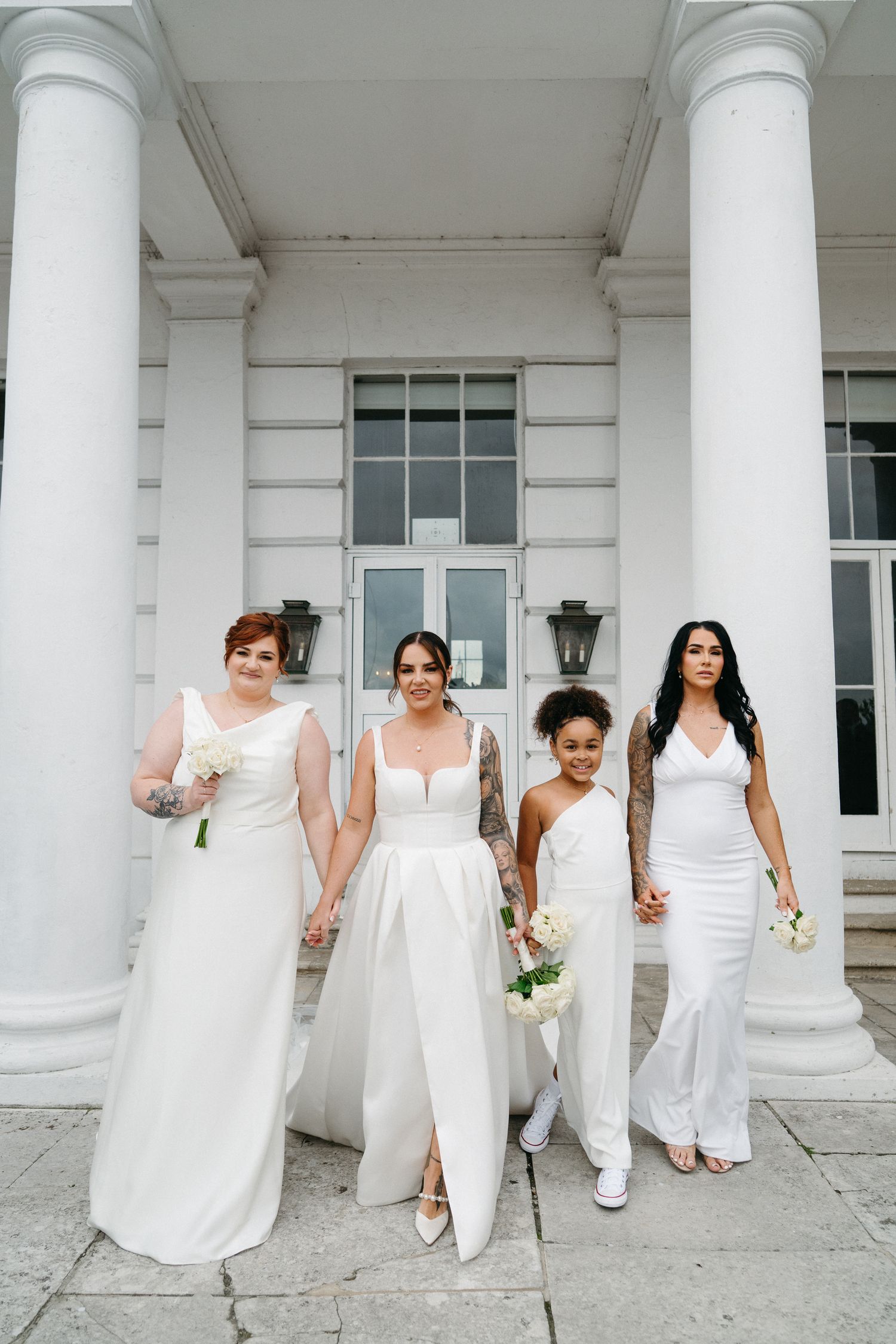 A group of bridesmaids in elegant white dresses pose together against a classic white columned building exterior.