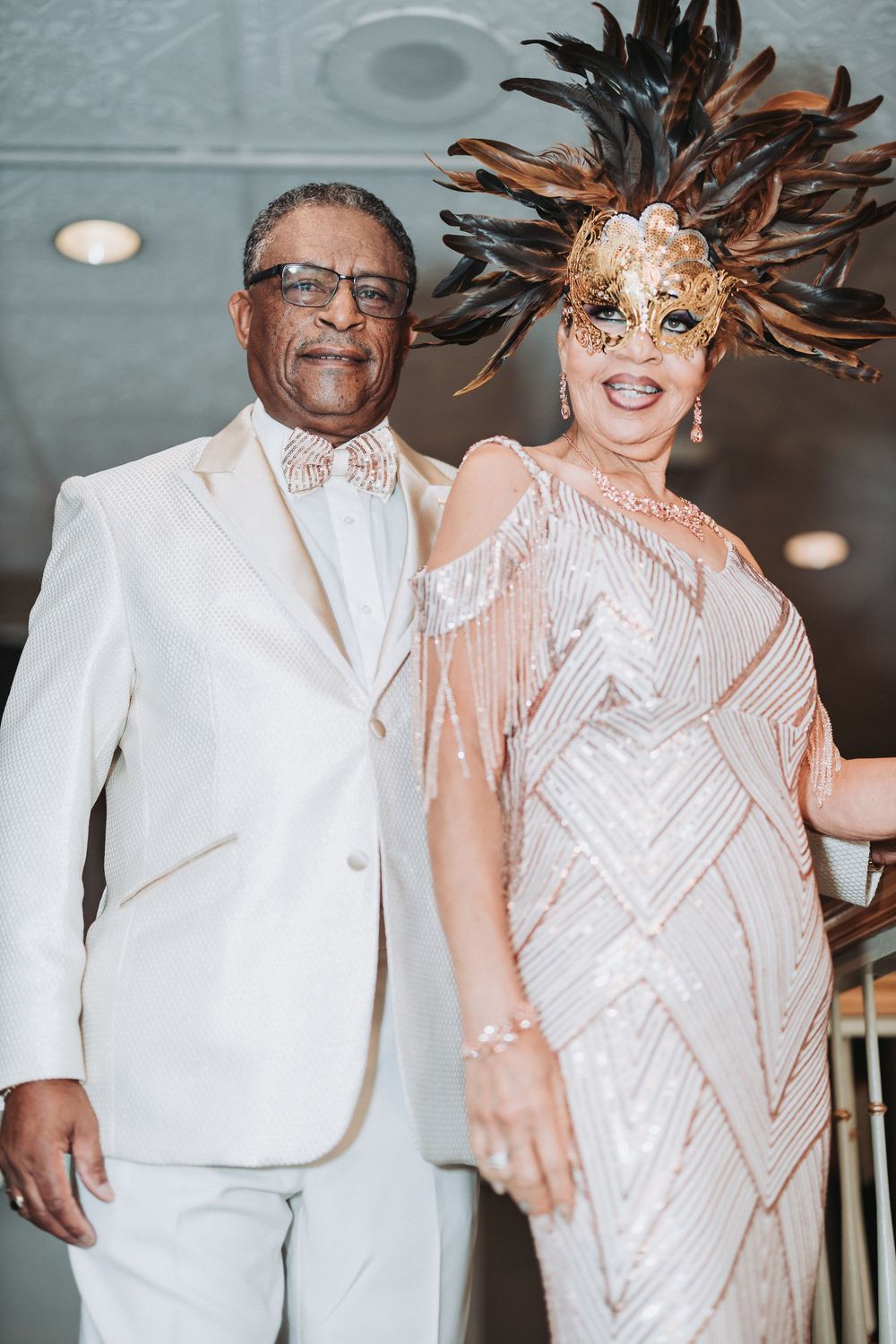 Two people at a formal event, one in a white suit and one in a sequined dress with feathered mask.