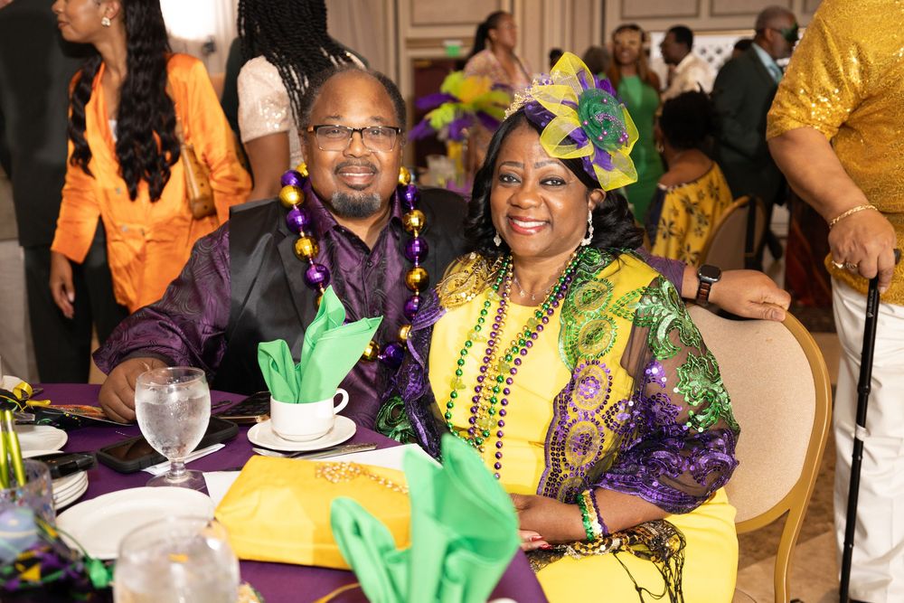 People in colorful Mardi Gras attire celebrate at a festive party with yellow and purple decorations.