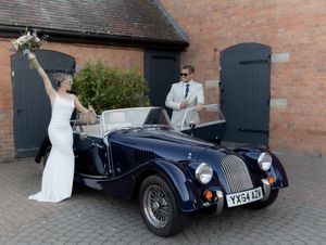 A classic navy Morgan sports car with a couple in wedding attire posing beside black doors of a brick building.