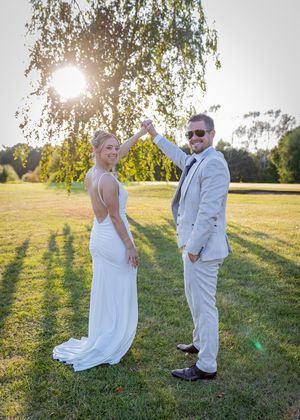 A couple in wedding attire poses in a sunlit meadow with a birch tree and golden sunlight creating long shadows.