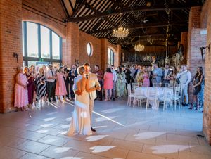 Wedding guests watch a couple share their first dance in a rustic brick barn venue with chandeliers overhead.