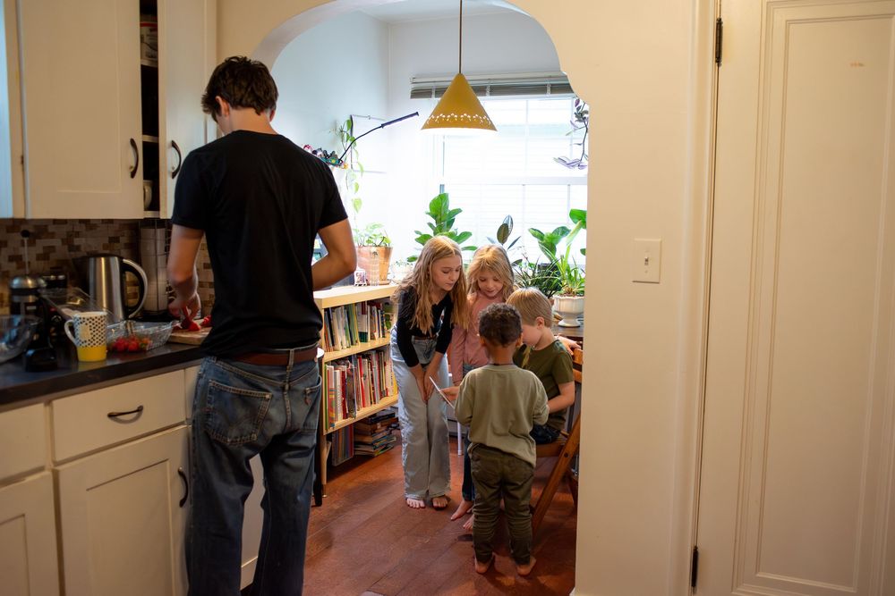 An oldest child making food while younger siblings look at a book in their family kitchen at home in Lansing, Michigan