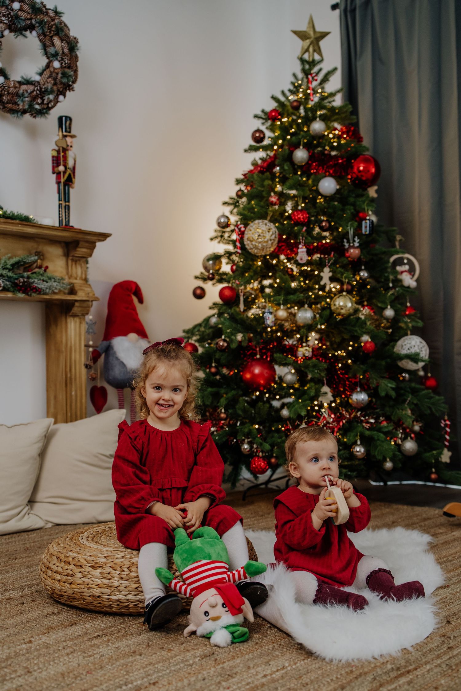 Children in red holiday outfits sit near decorated Christmas trees with ornaments and lights in a cozy living room setting.