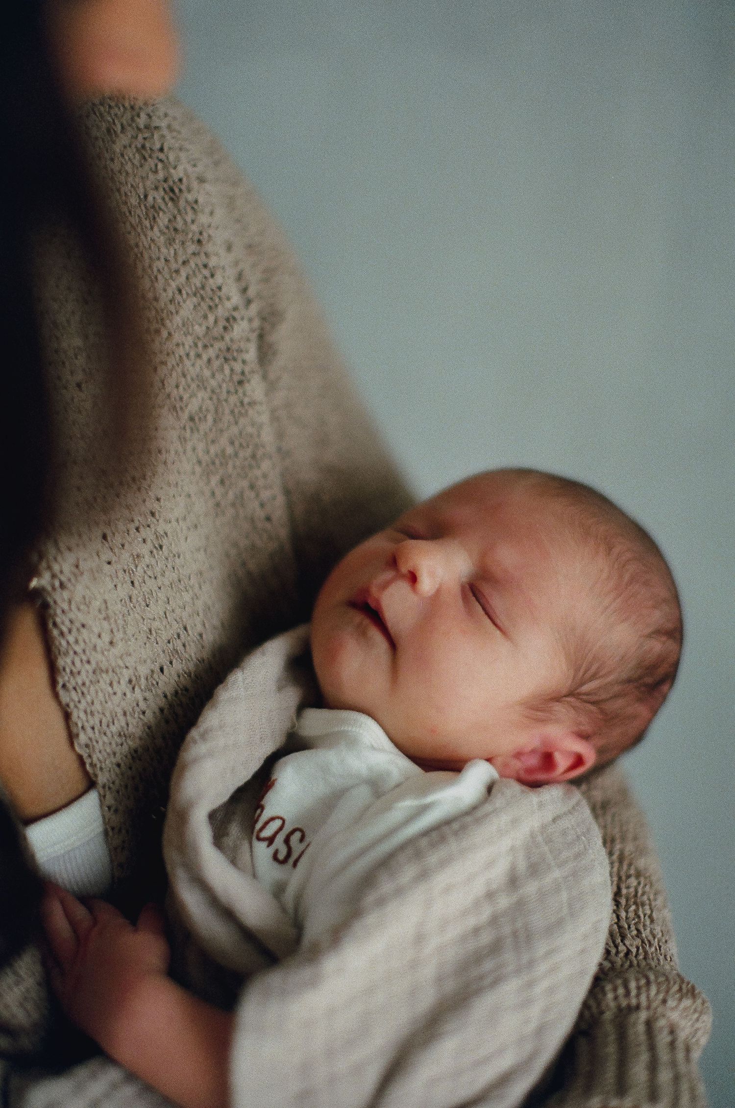 A sleeping newborn baby wrapped in a soft beige knit blanket rests peacefully against someone's shoulder.