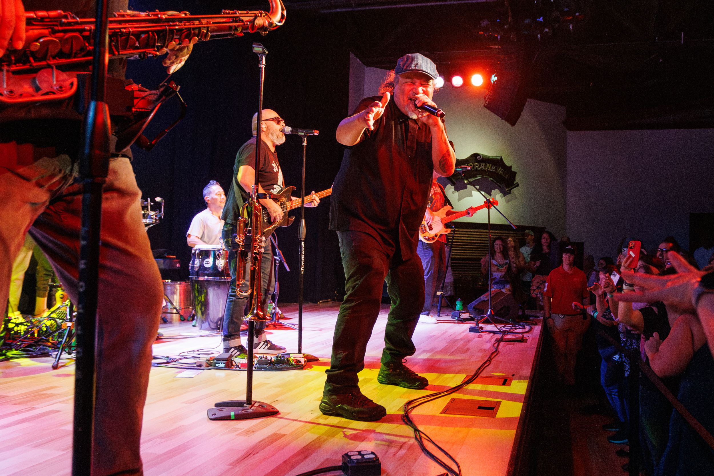 Musicians perform on stage at a concert venue with dramatic red lighting and music equipment visible in the background.