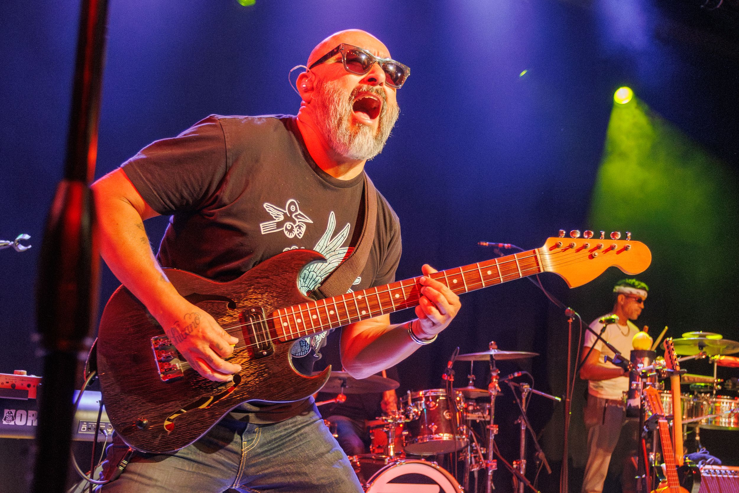 Musicians perform energetically on stage with electric guitars under dramatic blue and green lighting effects.
