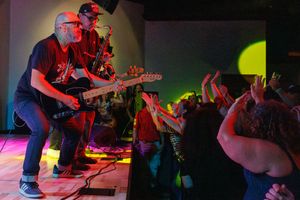 Musicians perform on stage with colorful lighting while audience members reach out toward them during a concert.