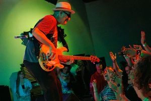 Live music performance featuring guitar player in orange shirt on stage with crowd silhouettes against green lighting effects.