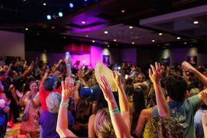 Concert crowd raises hands during performance with purple and pink stage lighting in a dark indoor venue.