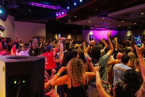 Concert attendees stand with arms raised in a dark venue with purple and blue lighting effects overhead.