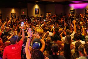 A large energetic crowd raises their hands during a concert with purple stage lighting illuminating the venue.