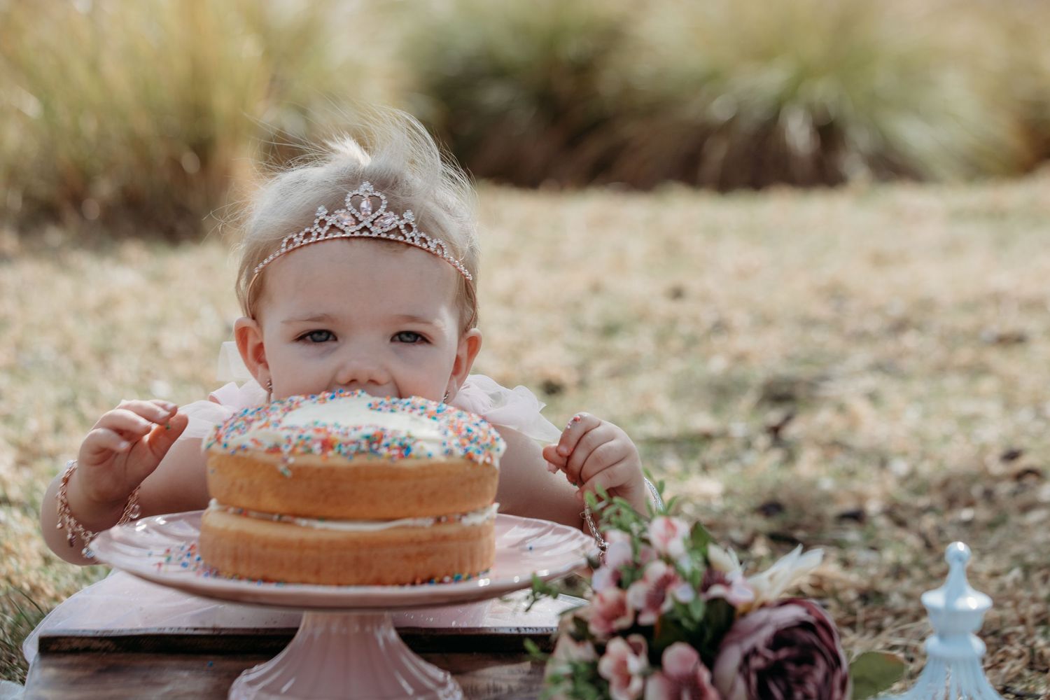 A colorful crown-shaped tiara sits atop a pink cake decorated with sprinkles and floral elements.
