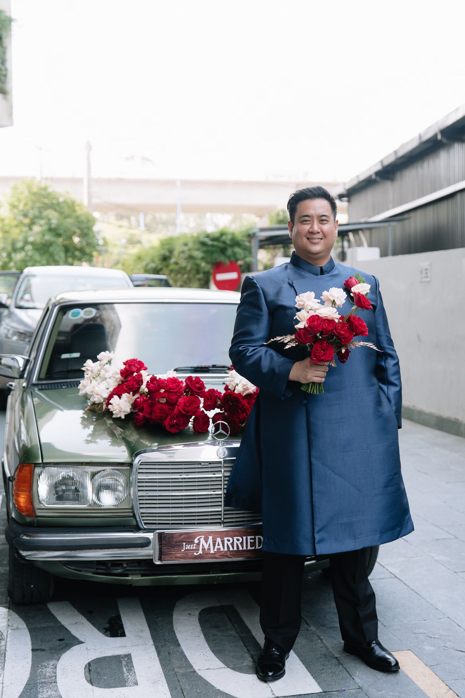 Groom in navy ao dai holding red roses beside a flower-decorated Mercedes Just Married wedding car.