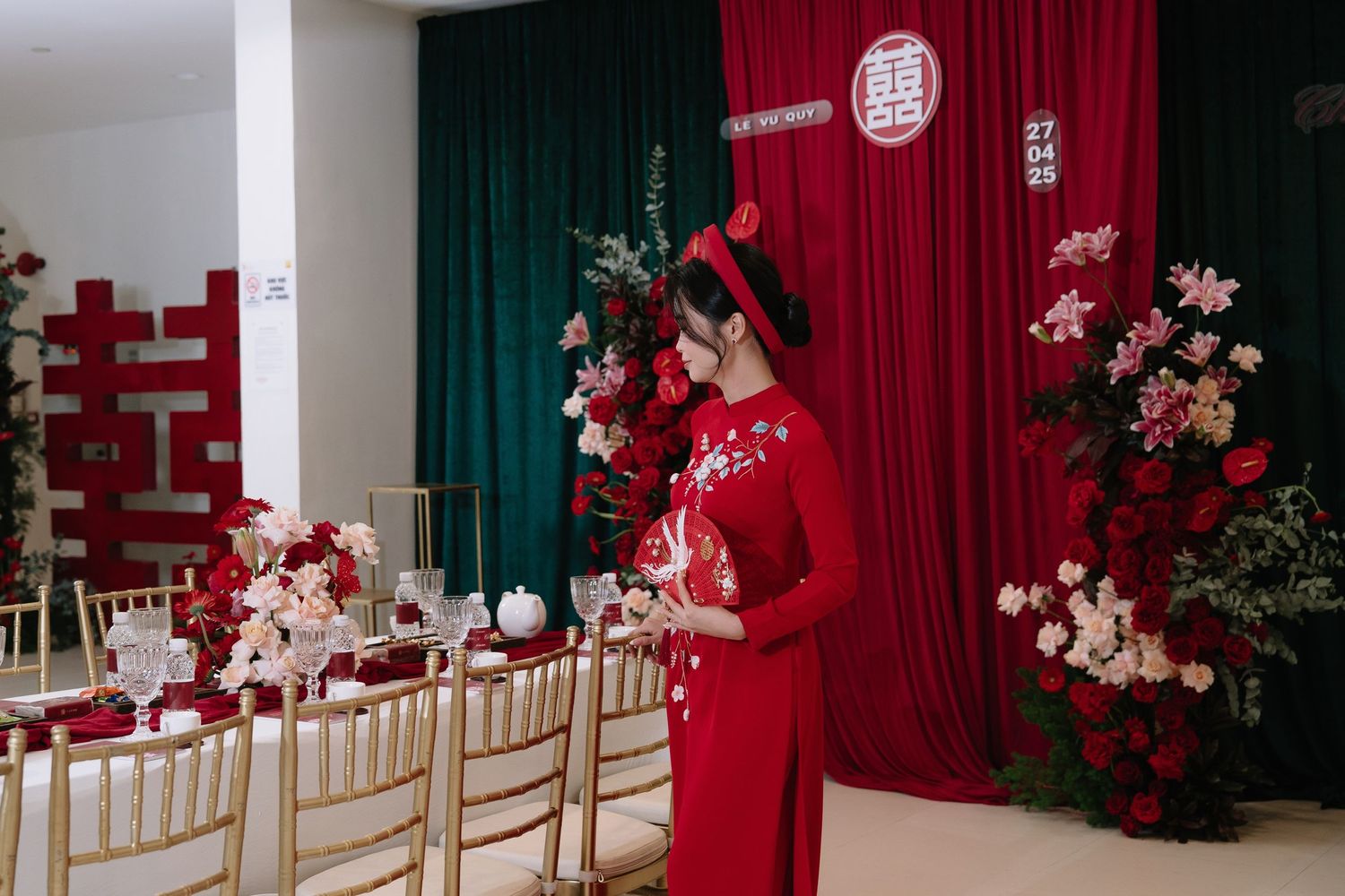 Vietnamese bride in red ao dai standing beside elegant wedding reception table setup.