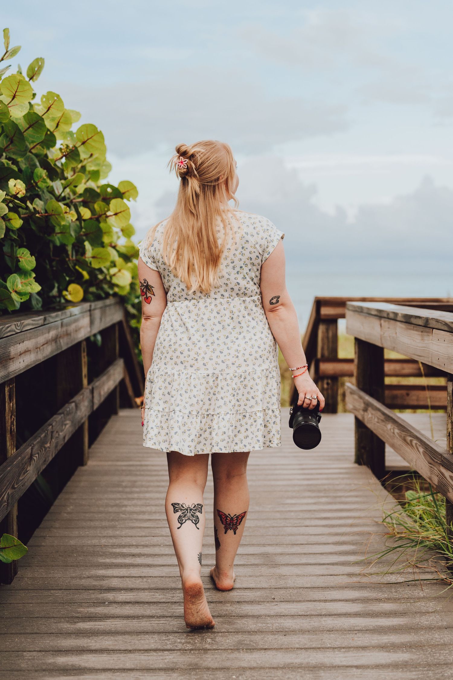 Woman in polka dot dress with butterfly leg tattoos walks on wooden boardwalk holding camera on sunny day.