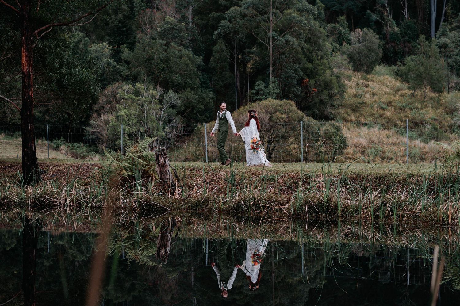 A couple stands on a wooden bridge over reflective lake waters surrounded by lush forest scenery.