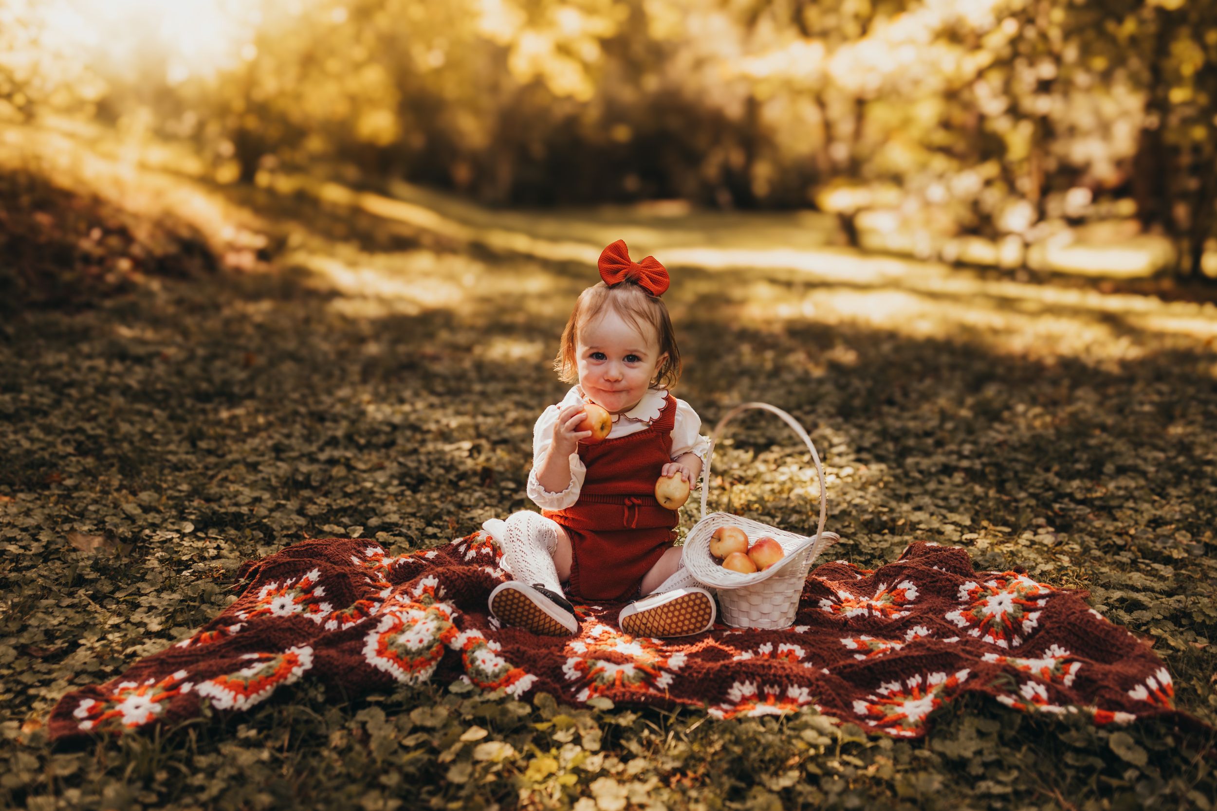 Baby girl in red romper sitting on vintage blanket with apple basket in warm golden autumn sunlight.