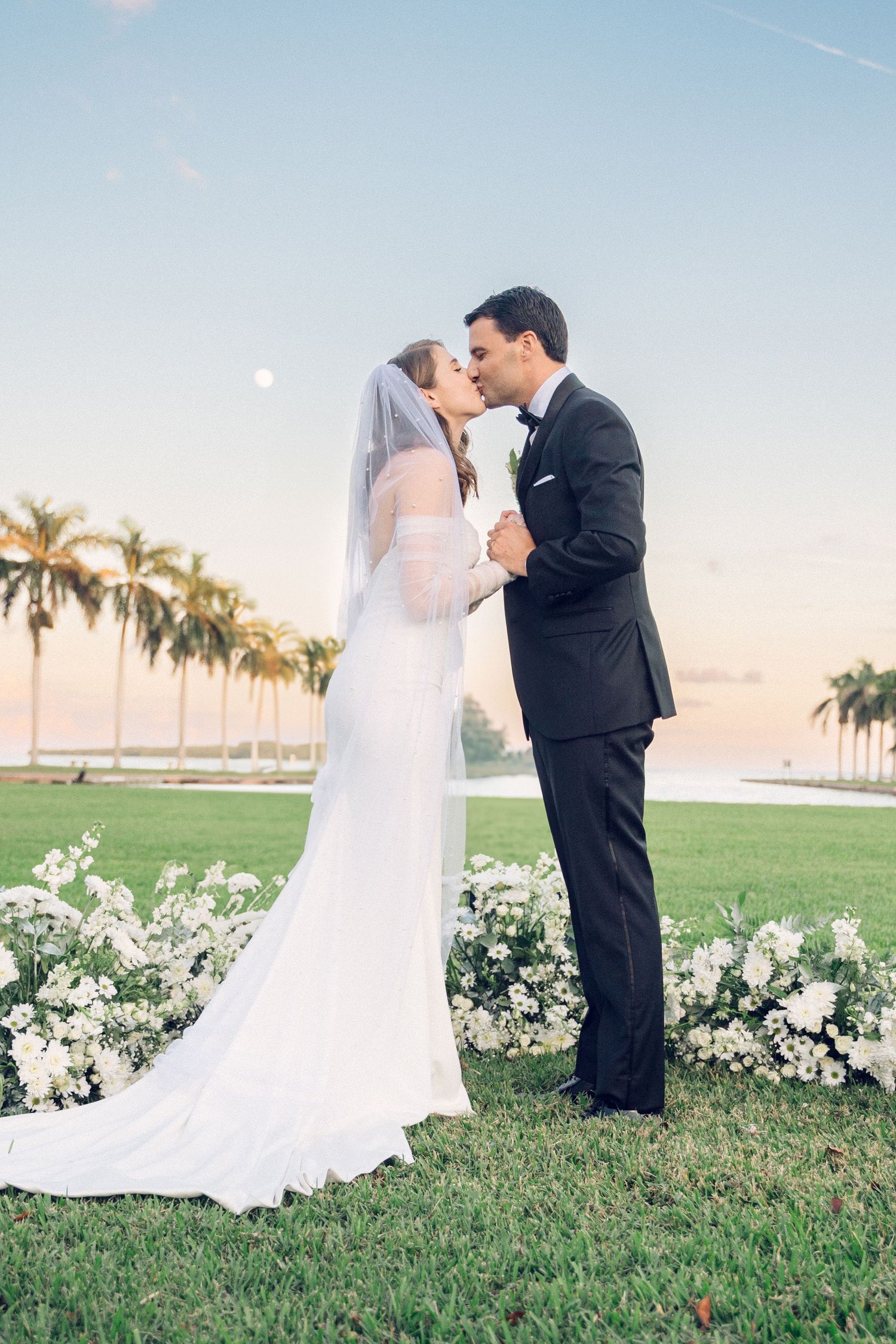 Romantic sunset wedding portrait with palm trees and white floral backdrop.