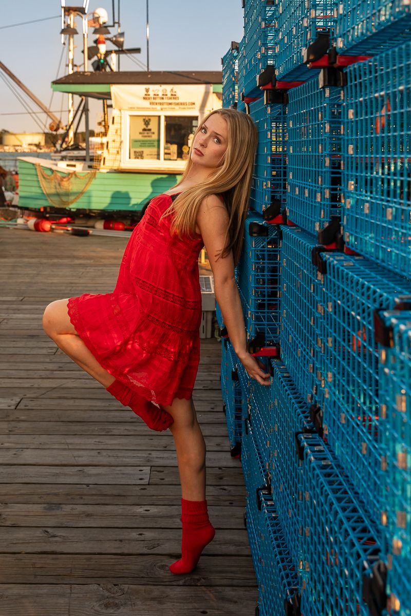 A person in a flowing red dress and socks poses playfully against blue lobster traps on a wooden dock at sunset.