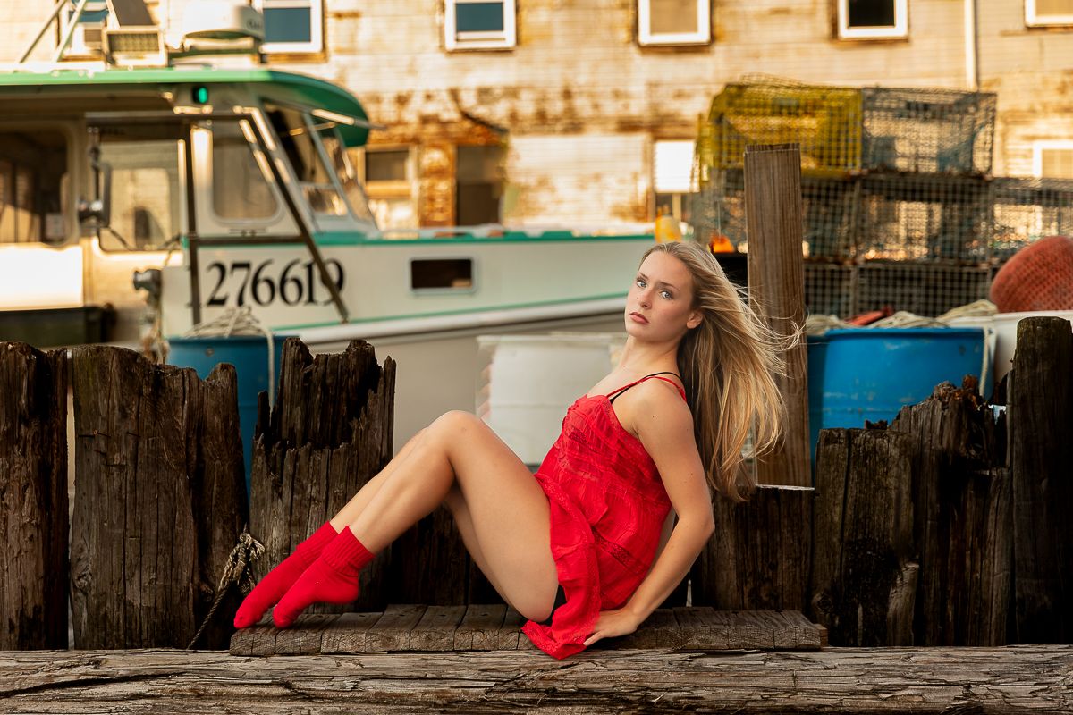 A person in a vibrant red dress and matching boots relaxes casually on a wooden dock at sunset.