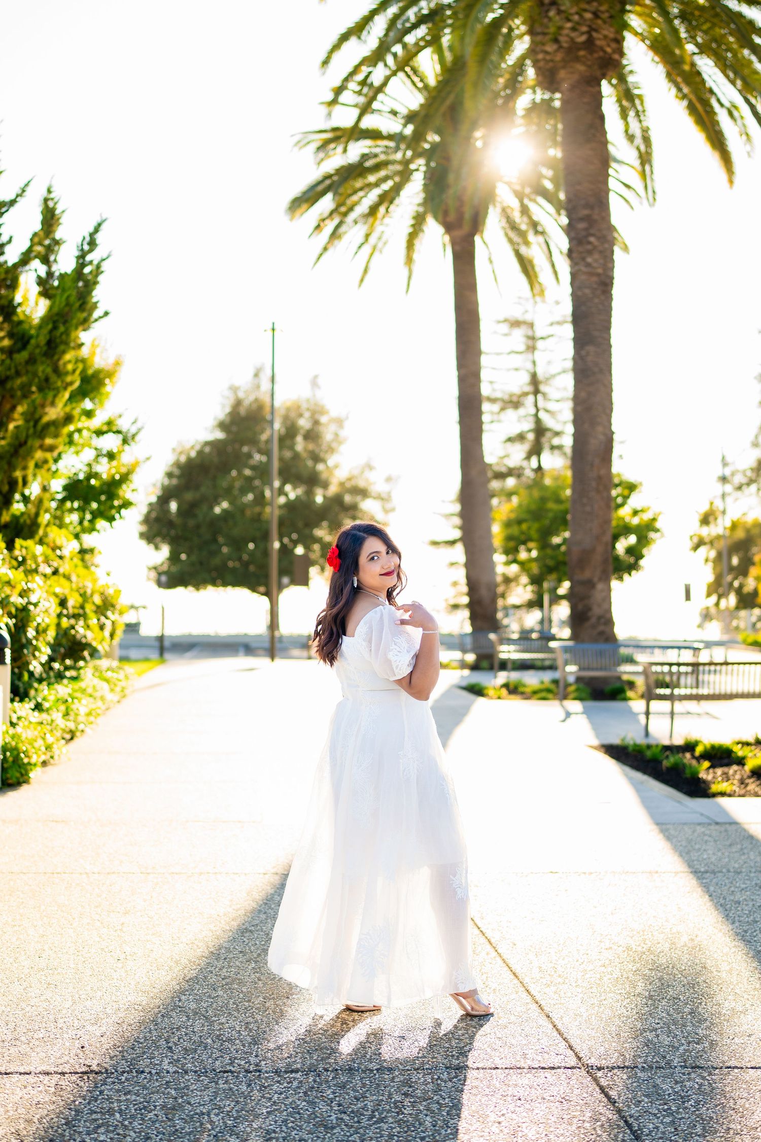 Woman in flowing white dress poses on sunlit path with palm trees and sunburst in tropical garden setting.