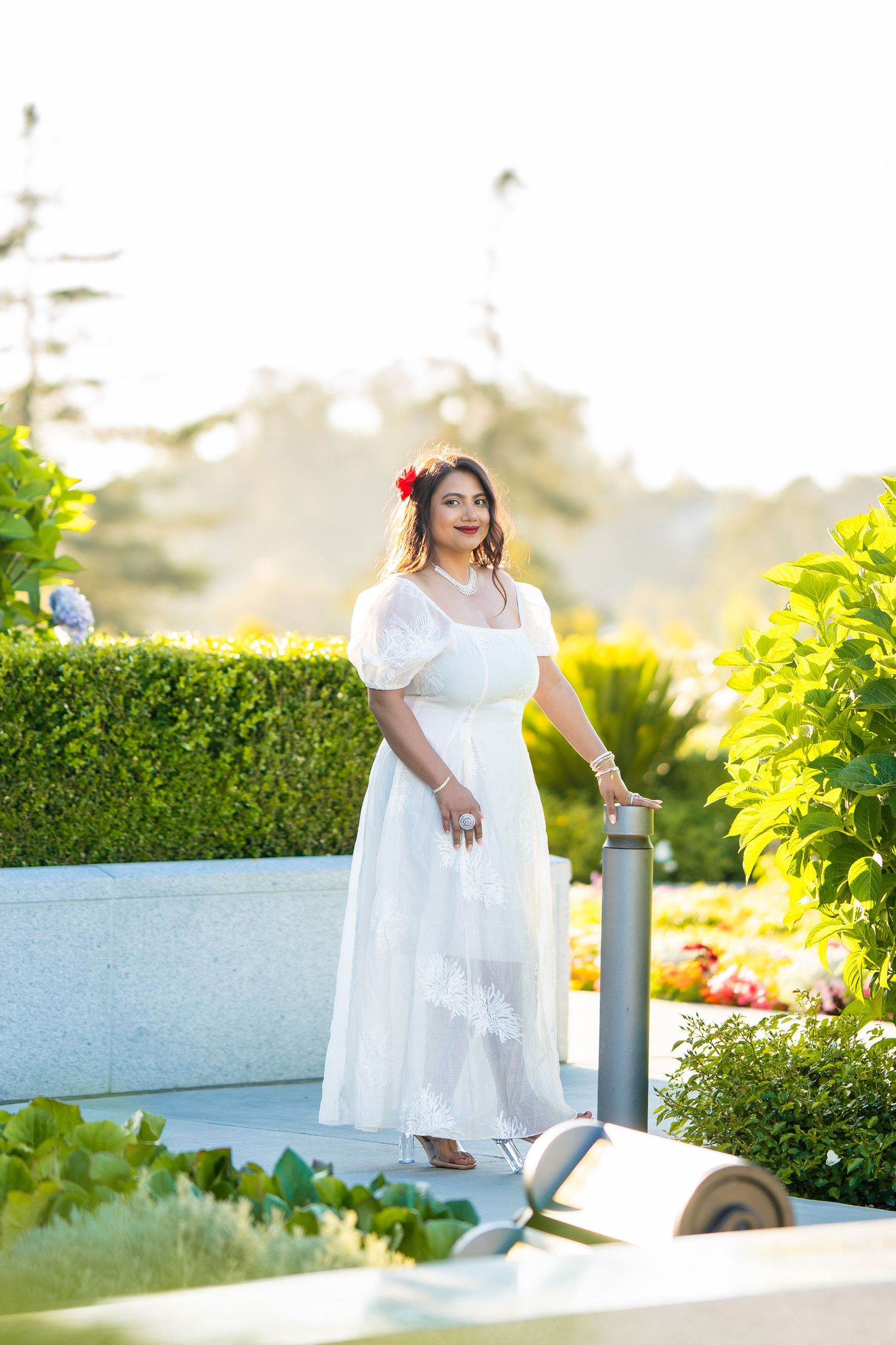 A woman stands in a white dress amid lush garden landscaping with palm trees and hedges at sunset.