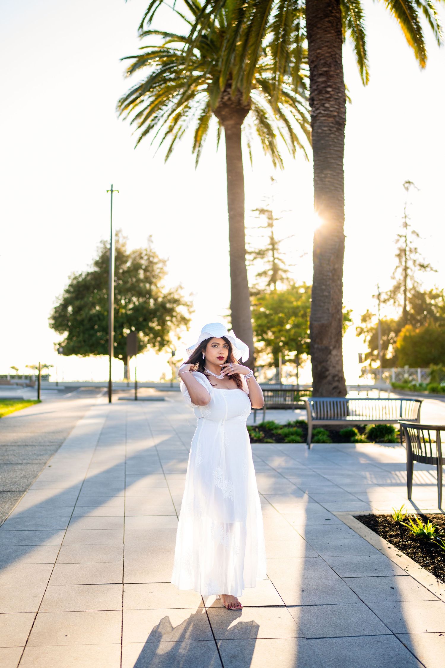 Woman in white dress dances joyfully between palm trees at sunset on paved walkway near waterfront.