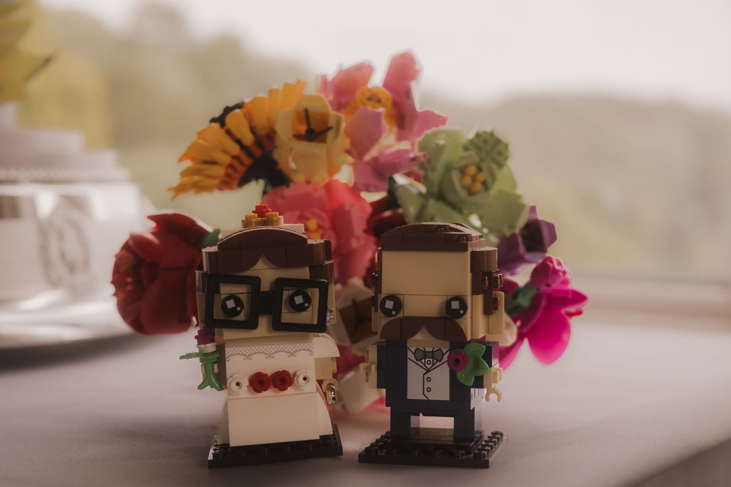 Two LEGO bride and groom figurines stand together with colorful flowers arranged behind them on a blurry surface.