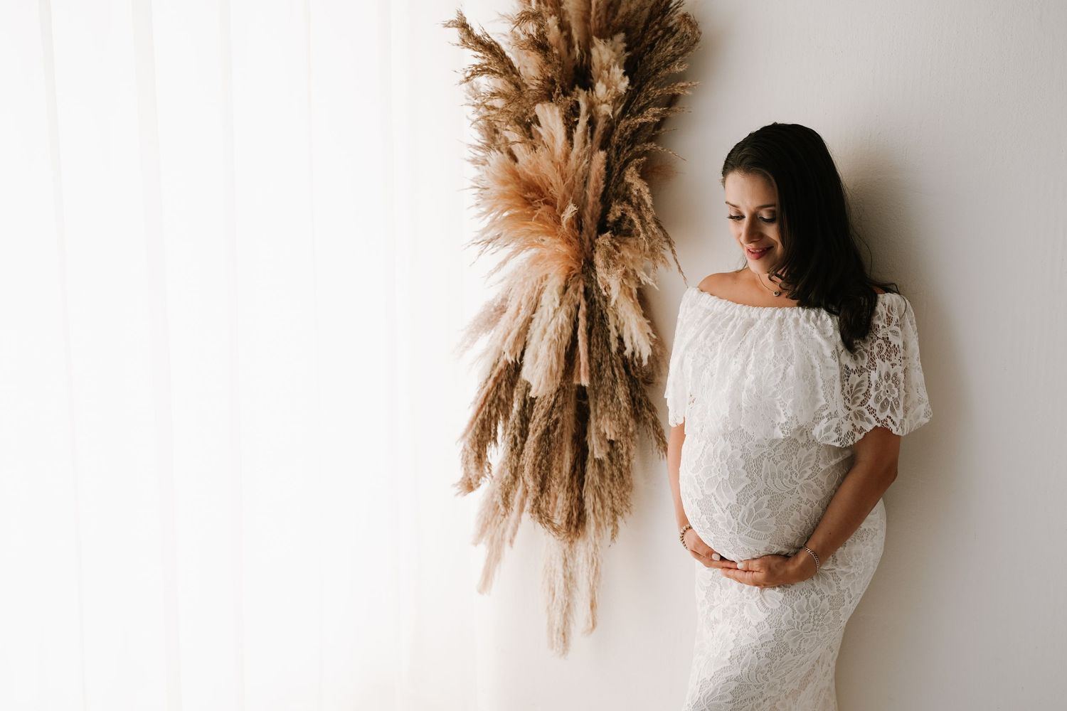 Pregnant woman in white lace dress cradling baby bump beside dried pampas grass wall decoration.
