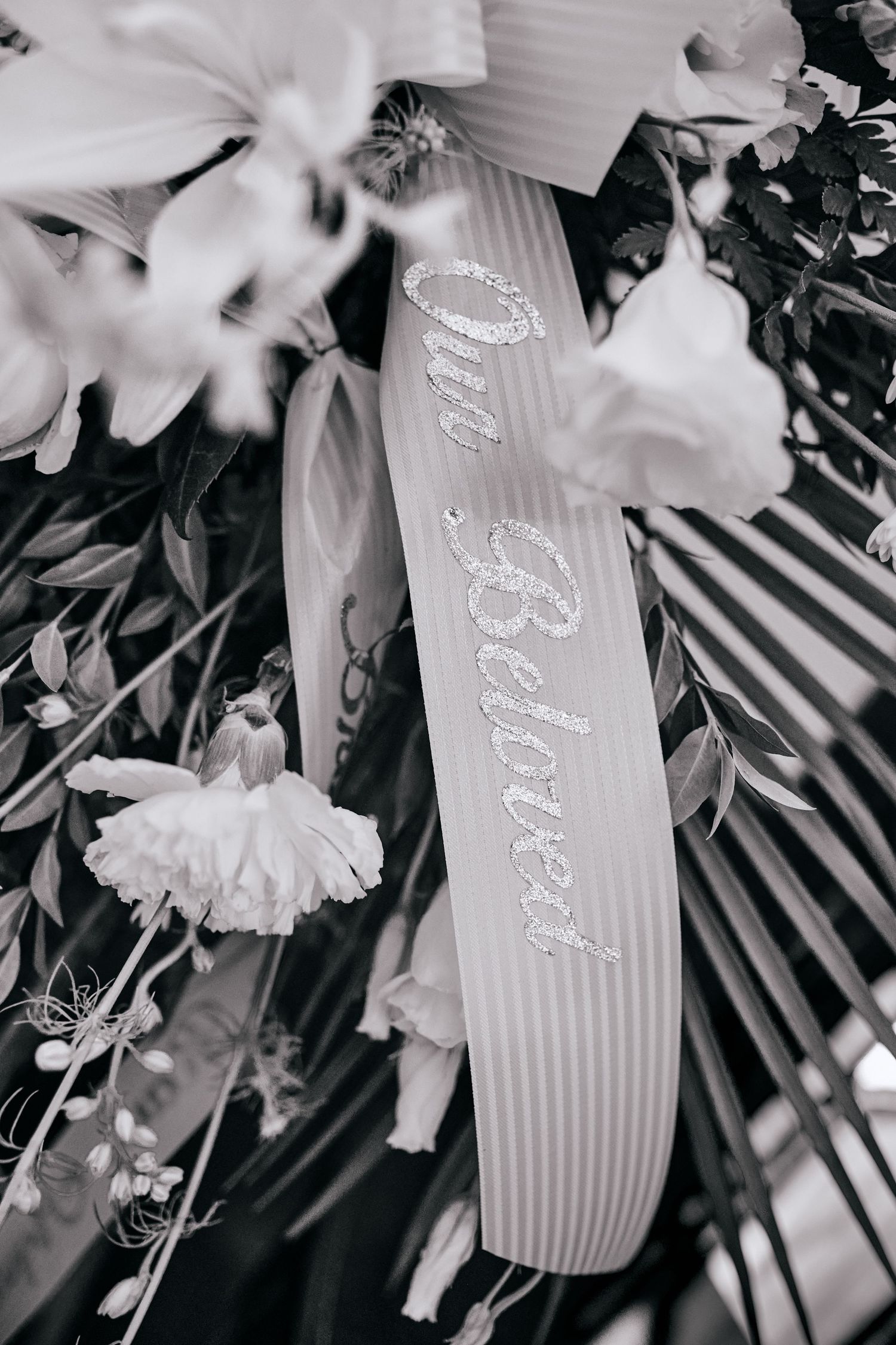 Black and white photo of a wedding sash with 'Mrs Balcom' text surrounded by white flowers and palm leaves.