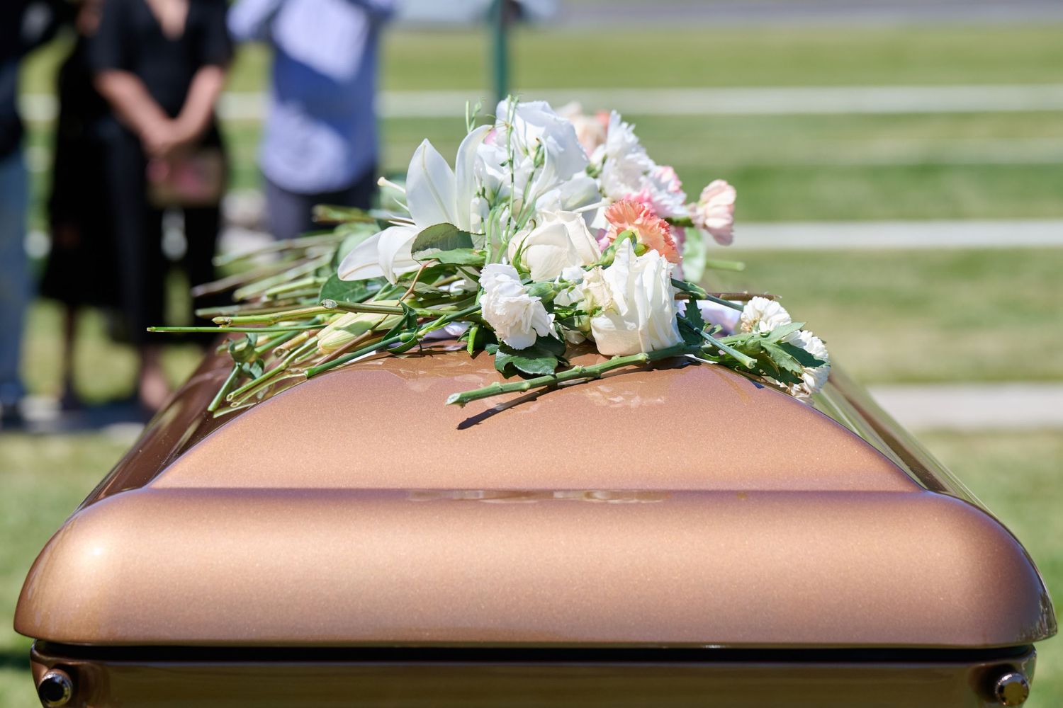 A delicate floral arrangement with white and peach flowers rests atop a rose gold colored casket at a funeral service.