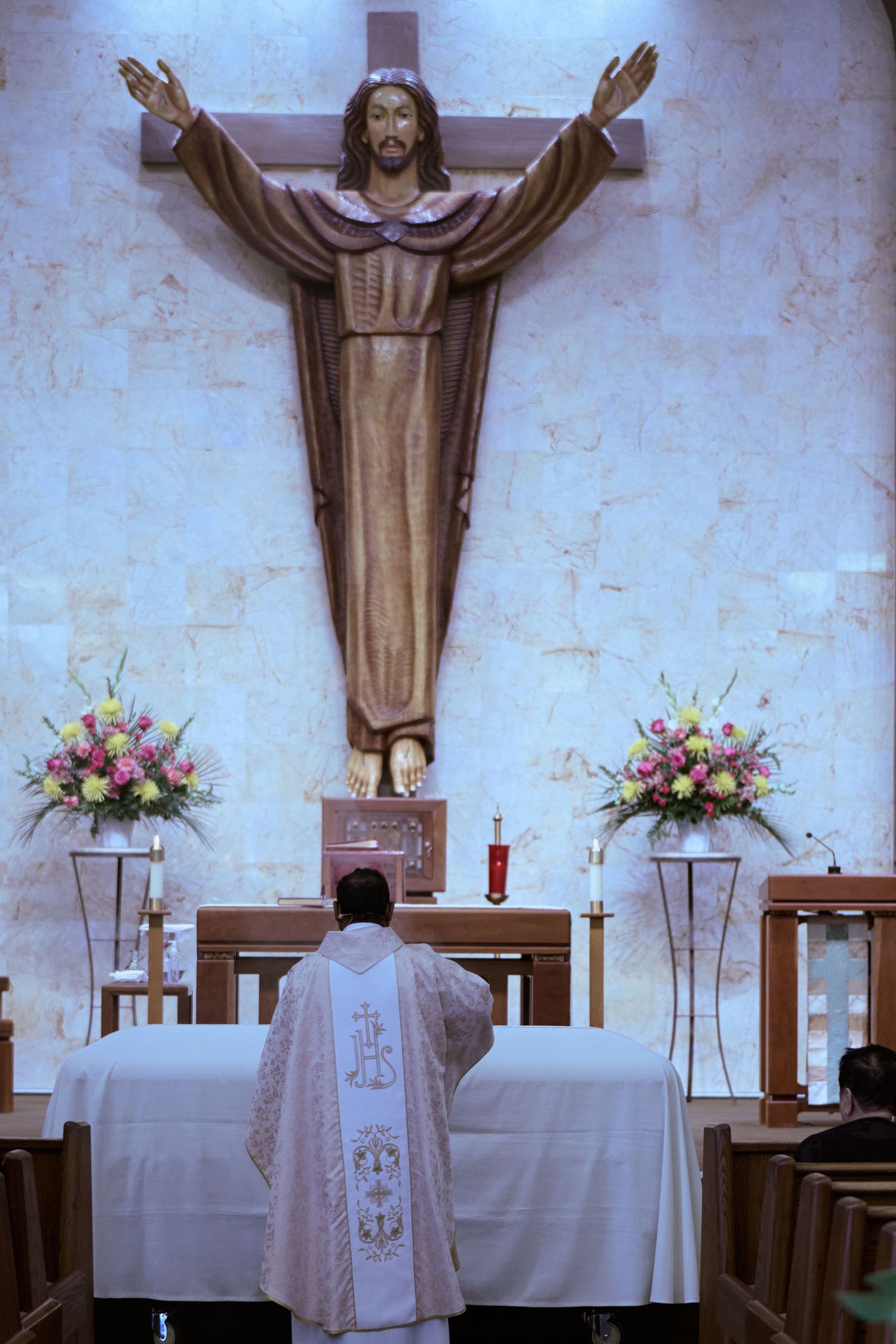 Religious altar in church with white cloths, pink flower arrangements, and large wooden crucifix figure mounted on wall.