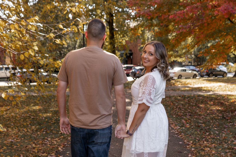 A couple walks through vibrant fall foliage on a sunny autumn day, surrounded by red and orange maple leaves.
