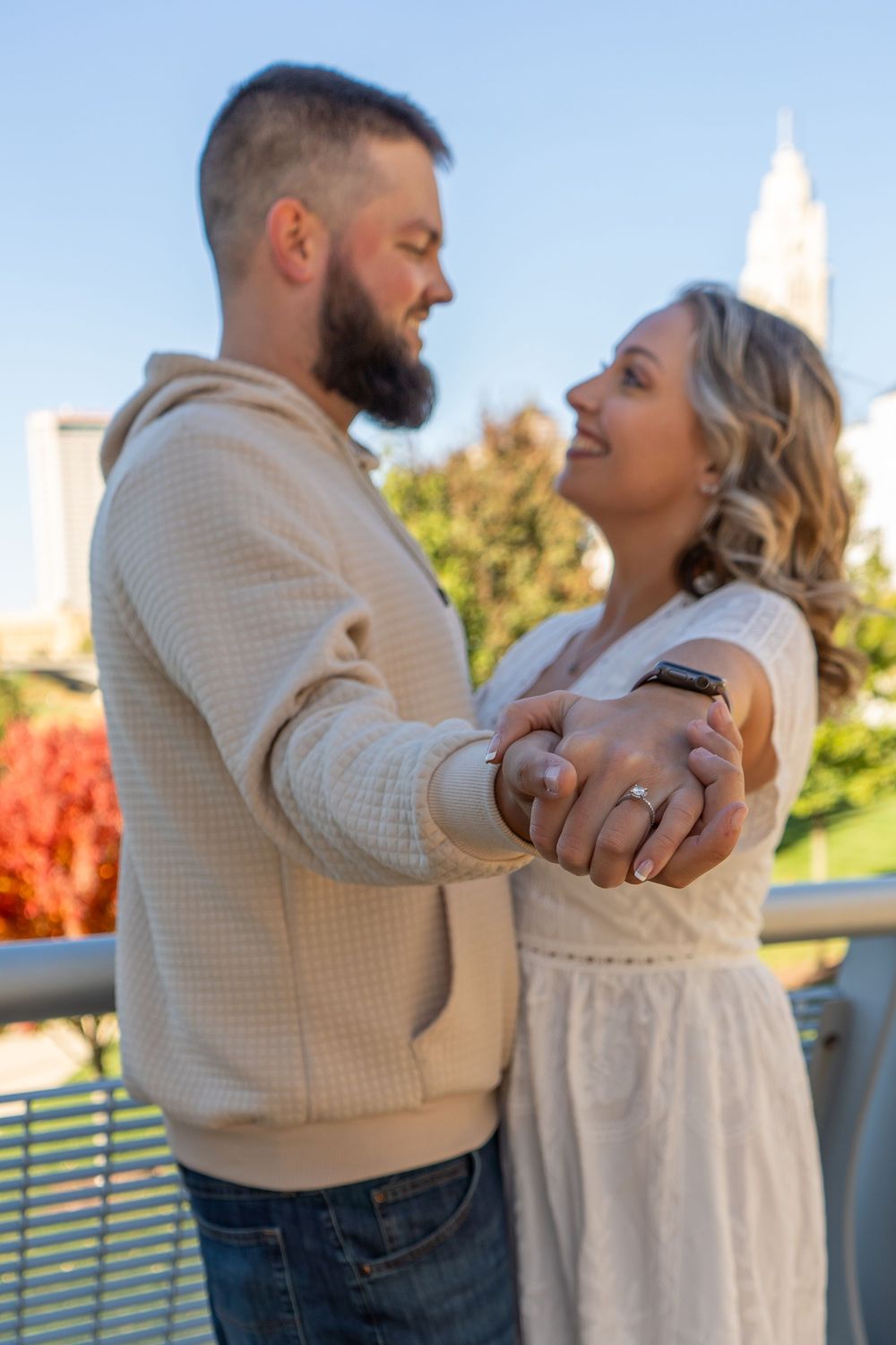 A couple in light colored clothing embrace outdoors on a sunny day with fall foliage and architecture visible in background.